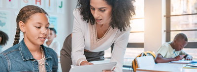 Eine Lehrerin erklärt einem Mädchen etwas auf dem Tablet. Sie steht bei der Schülerin am Schreibtisch in einem Klassenzimmer.