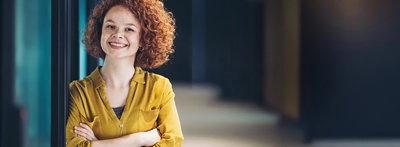 Rothaarige junge Frau mit gelber Bluse lehnt an Türrahmen und lächelt den Betrachter an.