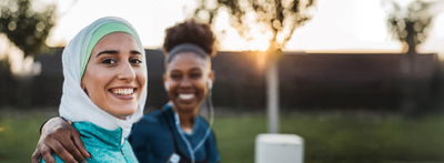 Zwei Frauen, eine mit Kopftuch und eine dunkelhäutig, lachen gemeinsam in die Kamera.