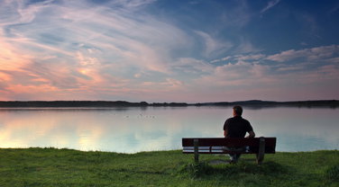 Ein Mann sitzt mirt Rücken zum Betrachter allein auf einer Bank an einem Seeufer.